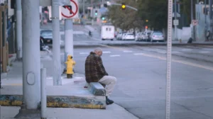 A man sitting alone on a curb beside a fire hydrant on a city street.
