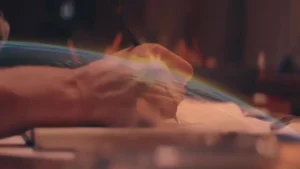 Close-up of a hand writing with a pen on paper by firelight, a rainbow flare across the frame.