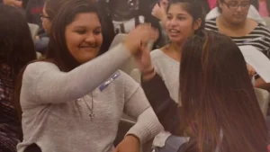 Teenage students in a classroom, one high-fiving another, both smiling.