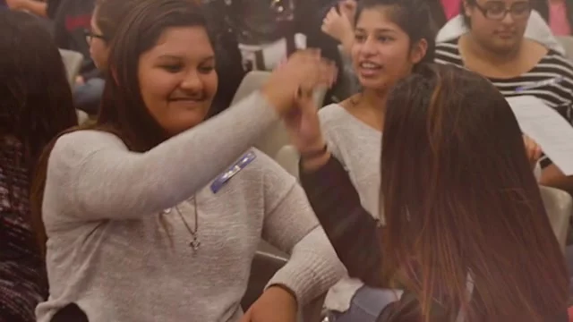 Teenage students in a classroom, one high-fiving another, both smiling.