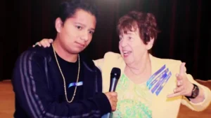 A woman speaking into a microphone on stage with her arm around a boy standing beside her.