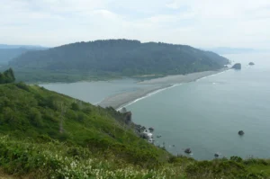 The mouth of the Klamath River meeting the Pacific, seen from a grassy headland above the beach.