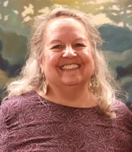 Robin Wall Kimmerer smiling, wearing a patterned plum top and long shell earrings, in front of a painted landscape backdrop.
