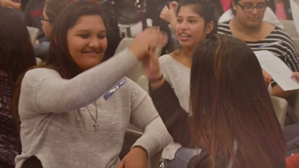 A high school student wearing a blue Who I Am Makes a Difference ribbon high-fives a classmate at a school assembly