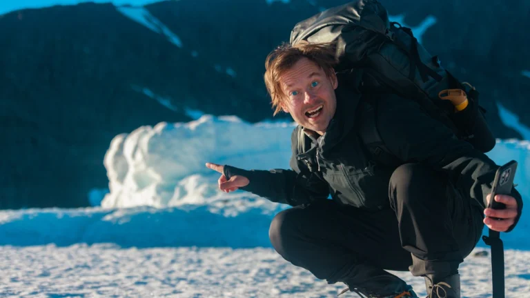 A person in outdoor clothing crouches on snow beside a glacier and points at the ice.