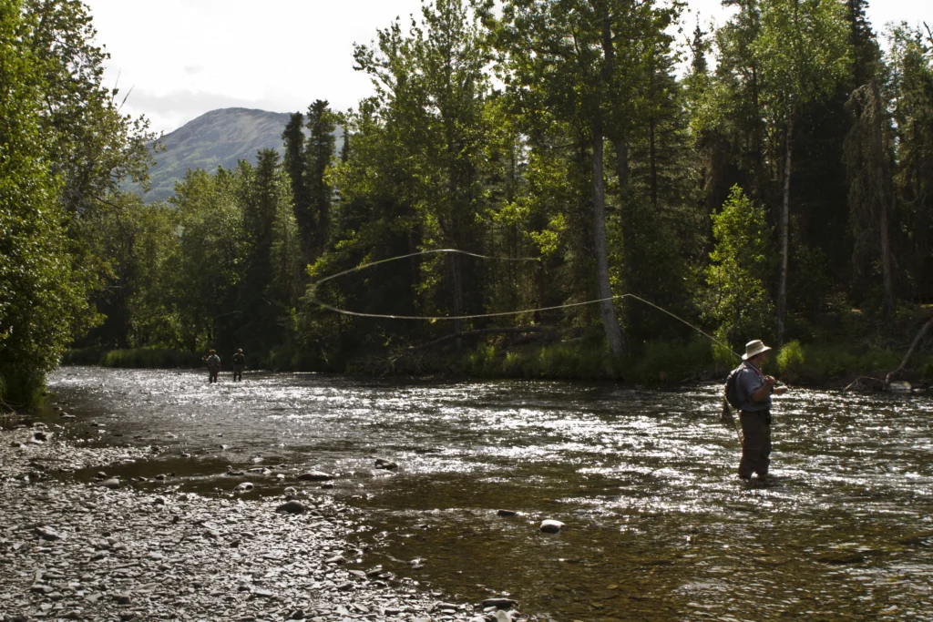 An angler casting a fly line in a wide river, with two more figures far upstream and forest on both banks. No face is readable. It illustrates fly fishing as a subject and does not show this show's hosts or guests.