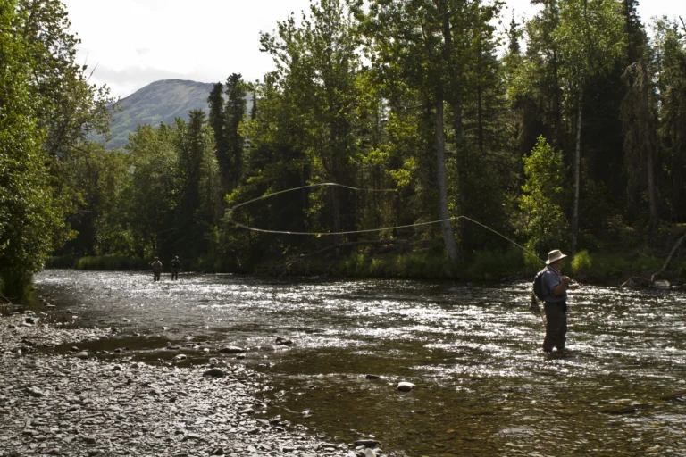 An angler casting a fly line in a wide river, with two more figures far upstream and forest on both banks. No face is readable. It illustrates fly fishing as a subject and does not show this show's hosts or guests.