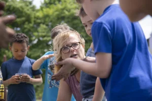 Illustration only, not a Blue Ribbons Worldwide program. A teacher crouches outdoors to listen as an elementary student shows her something in his hand, at a school in Gasport, New York, in 2022.
